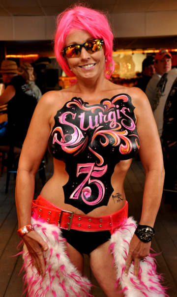woman with pink hair with a sturgis 75 body painting on her chest that is mostly pink on a black background